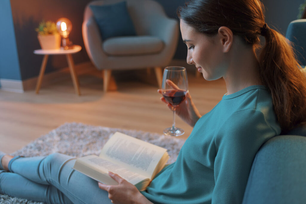 Young woman relaxing at home: she is sitting on the floor in the living room, drinking a glass of wine and reading a book